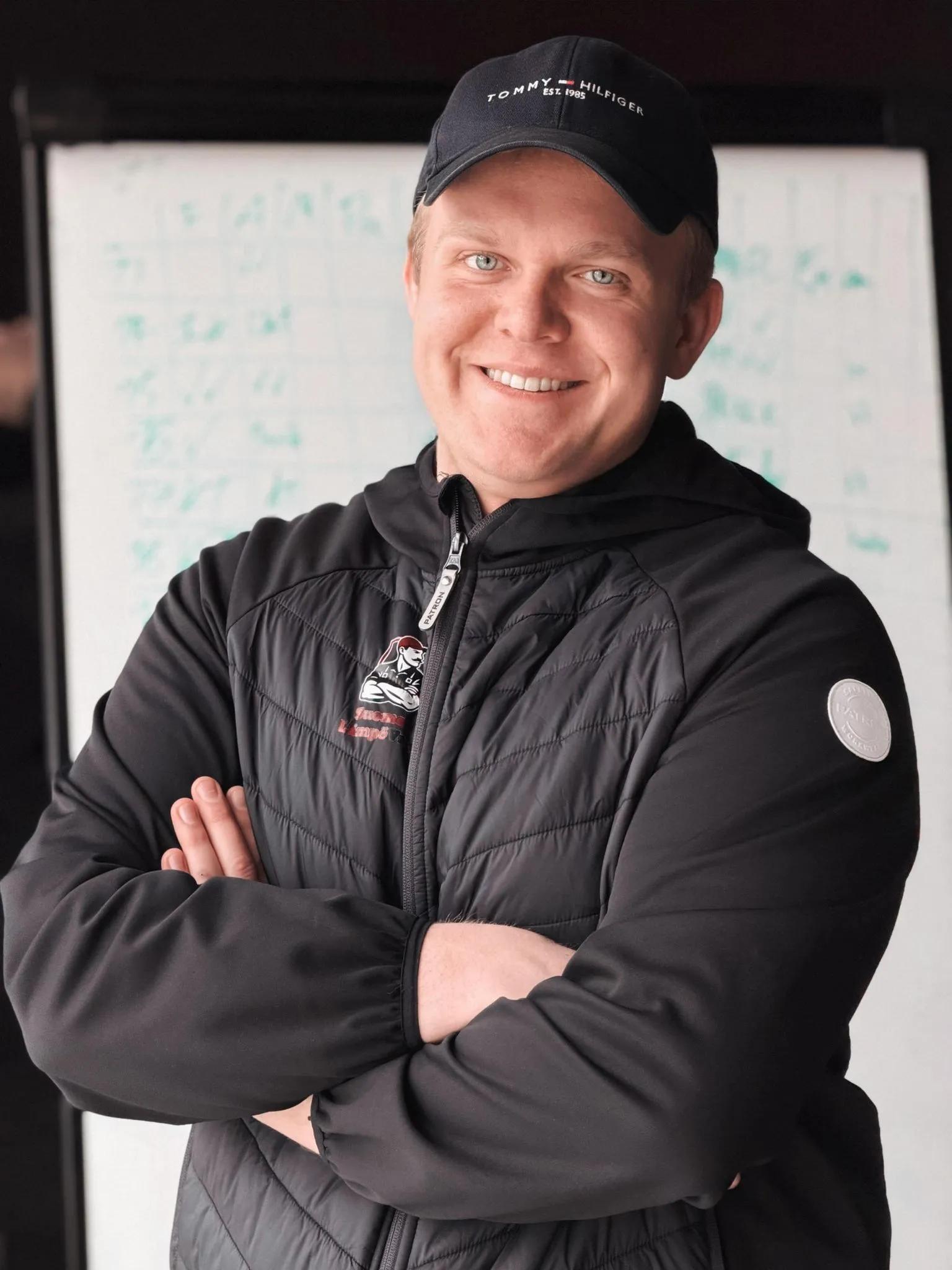 Hymyilevä mies seisoo kädet ristissä, yllään musta Tommy Hilfiger -lippis ja musta takki, jonka vasemmassa rinnassa on punainen logo. Taustalla näkyy valkotaulu, jossa on vihreää tekstiä. Valo on pehmeää ja tasaista.