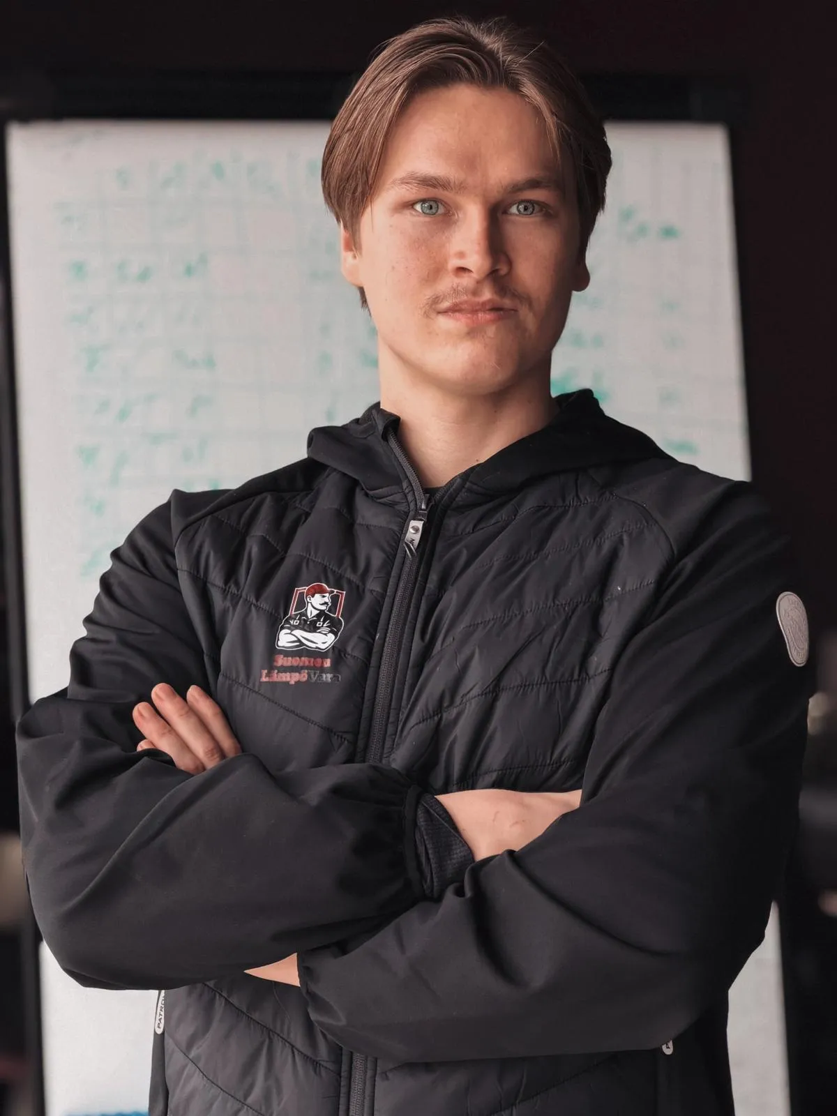 Muotokuva nuoresta miehestä, jolla on vaaleat hiukset ja siniset silmät, mustassa takissa, kädet ristissä, taustalla valkotaulu.