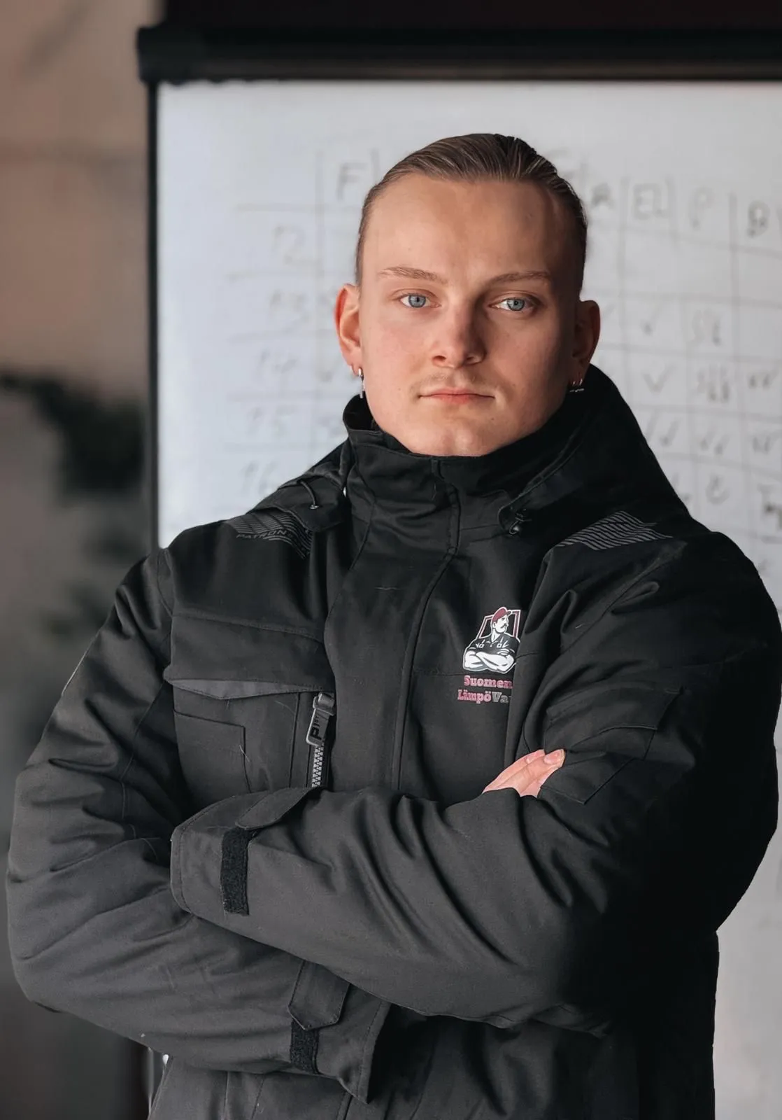 Muotokuva nuoresta miehestä mustassa takissa, kädet ristissä, taustalla valkotaulu.