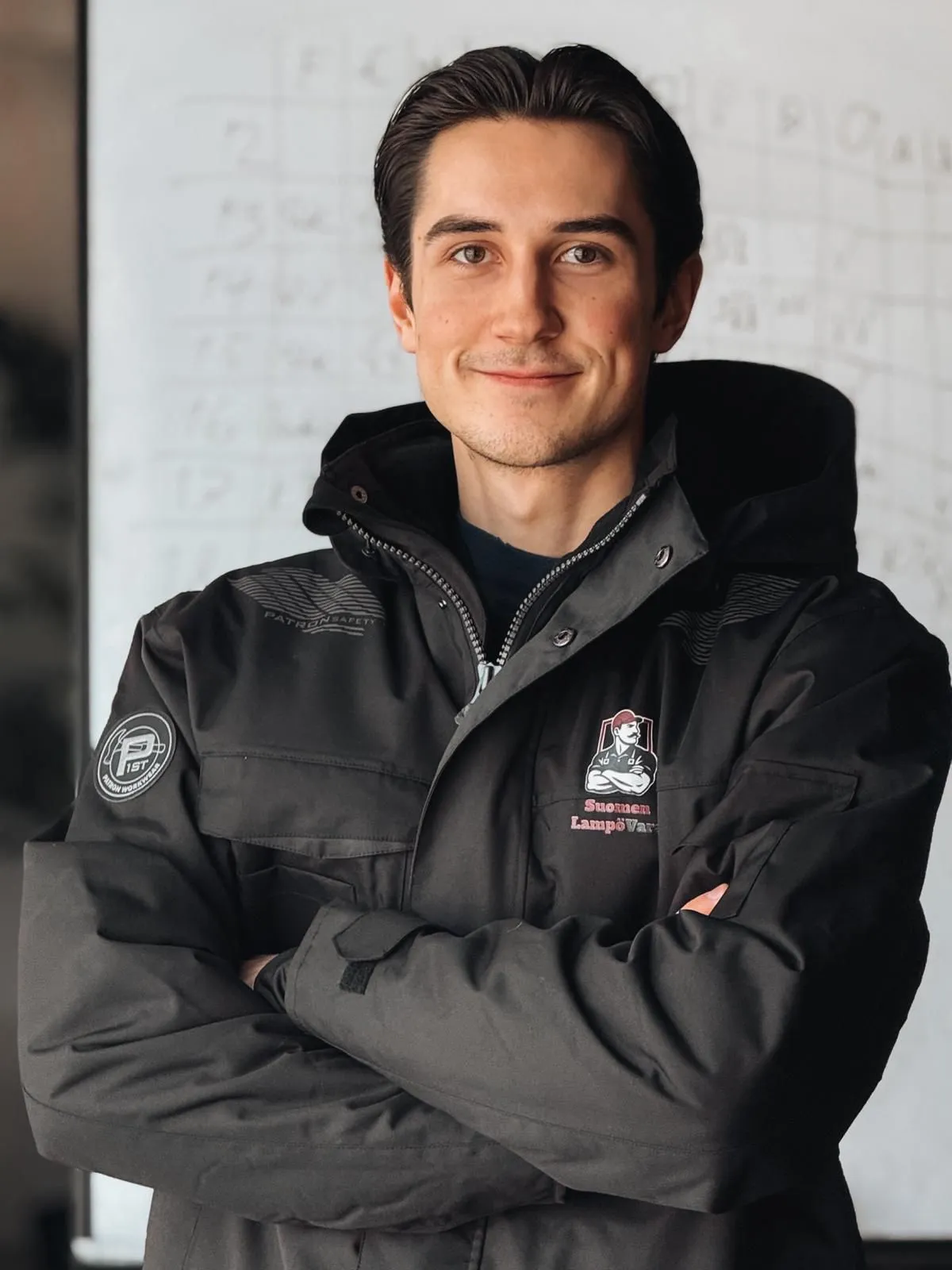 Muotokuva nuoresta miehestä mustassa takissa, jossa on logot ja tekstiä, valkoisen taulun edessä.