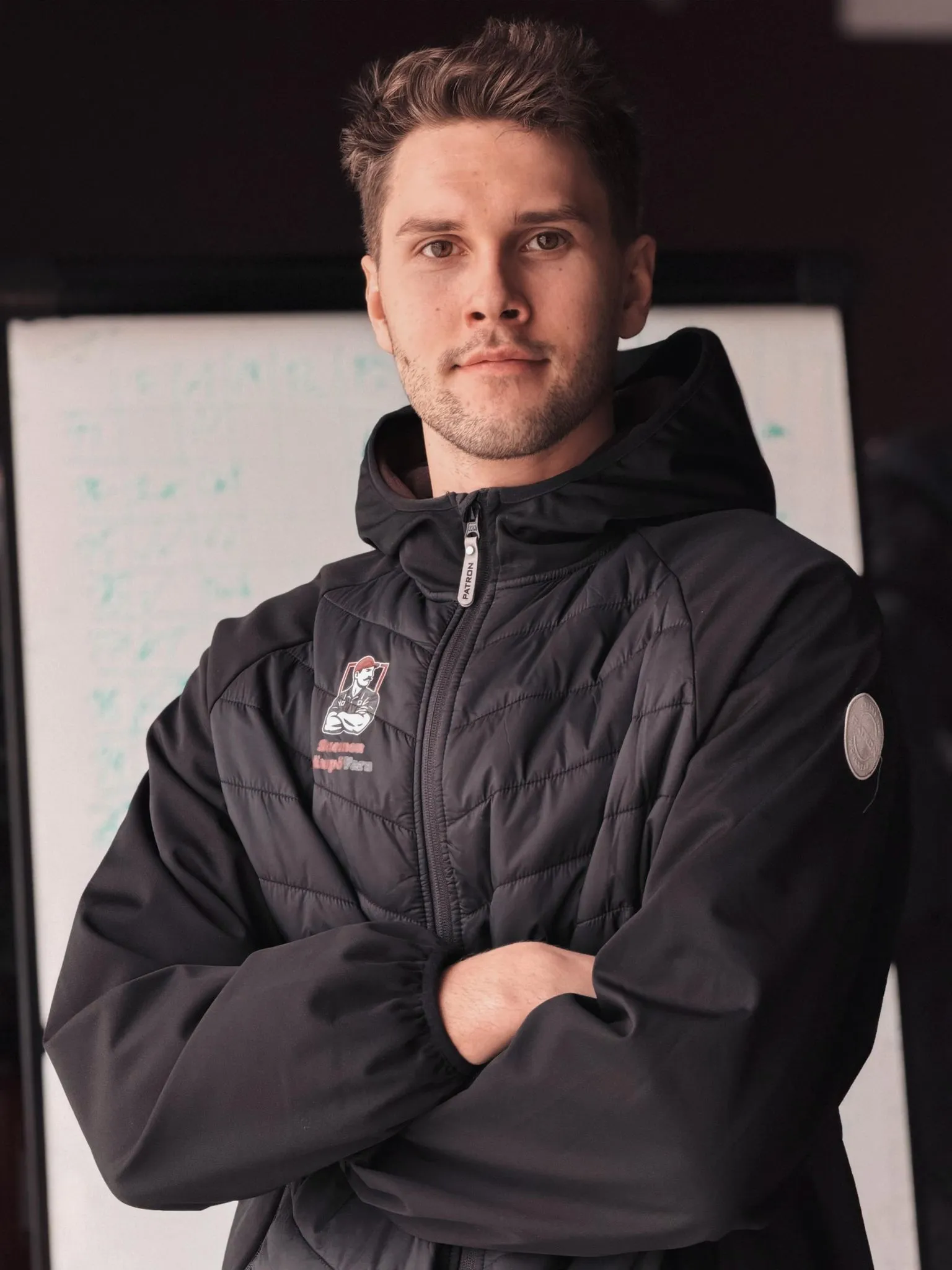 Muotokuva nuoresta miehestä mustassa toppatakissa, kädet ristissä, valkotaulun edessä.