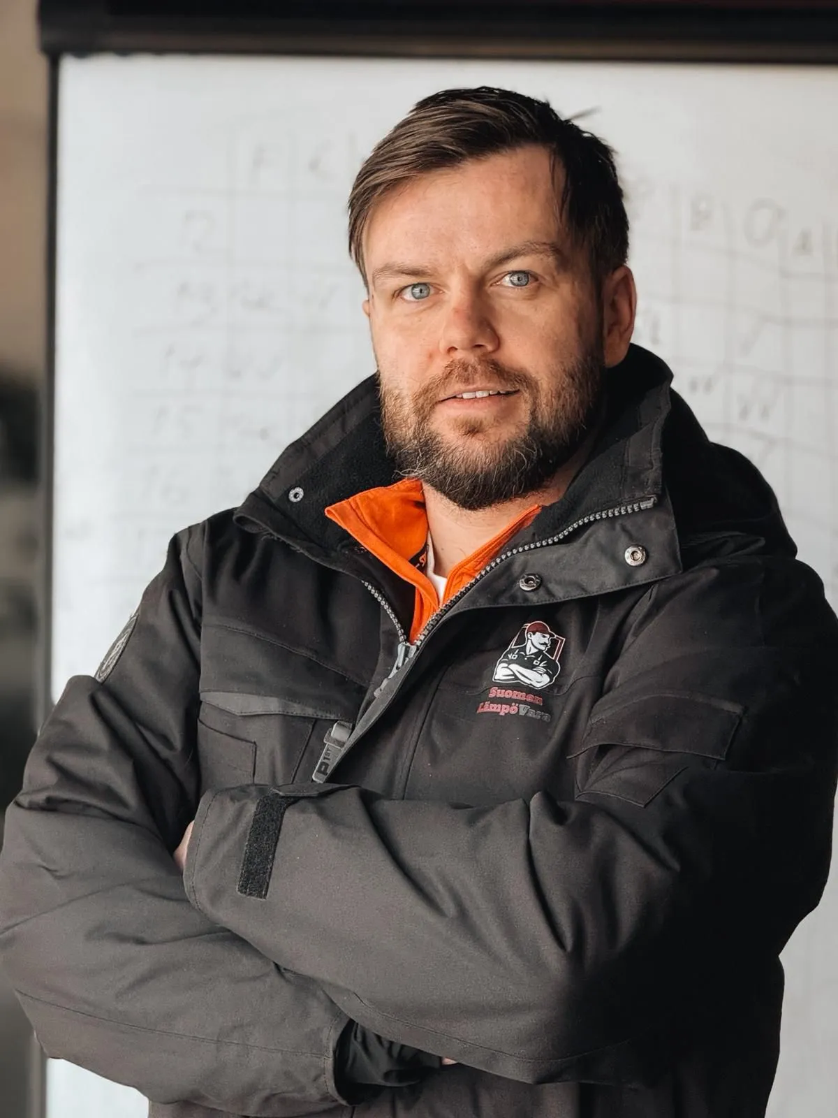 Muotokuva miehestä mustassa takissa, jossa on oranssi kaulus, kädet ristissä ja taustalla valkotaulu.