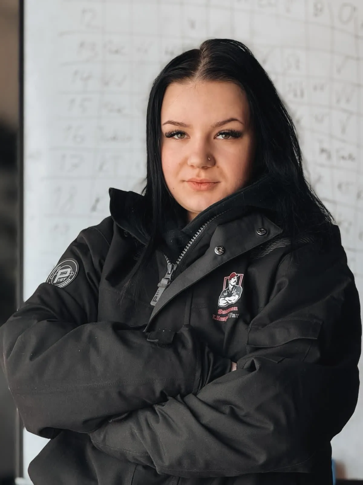 Nuori nainen mustassa takissa seisoo kädet ristissä, taustalla valkotaulu, jossa on tekstiä.