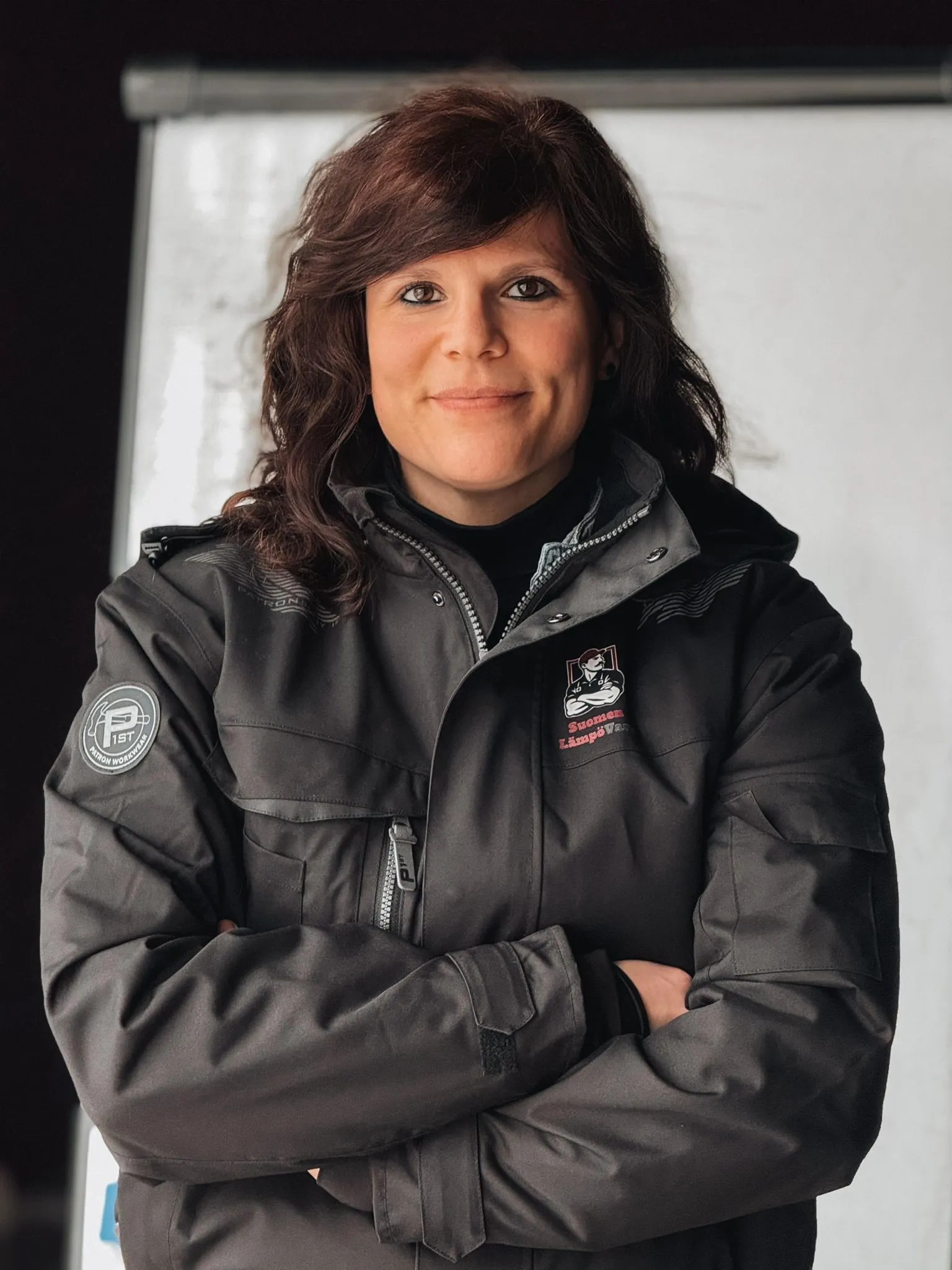 Muotokuva naisesta, jolla on ruskeat hiukset ja yllään tumma takki, jossa on yrityksen logot, valkoisen taustan edessä.